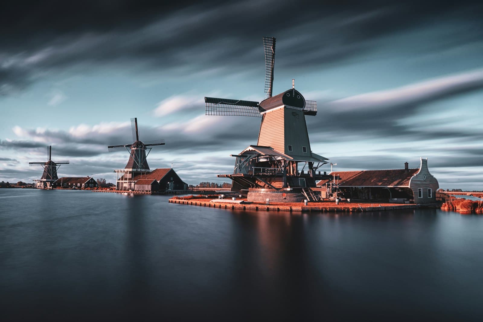 Drie houten windmolens aan stil water onder snel bewegende wolken, gefotografeerd met een lange sluitertijd